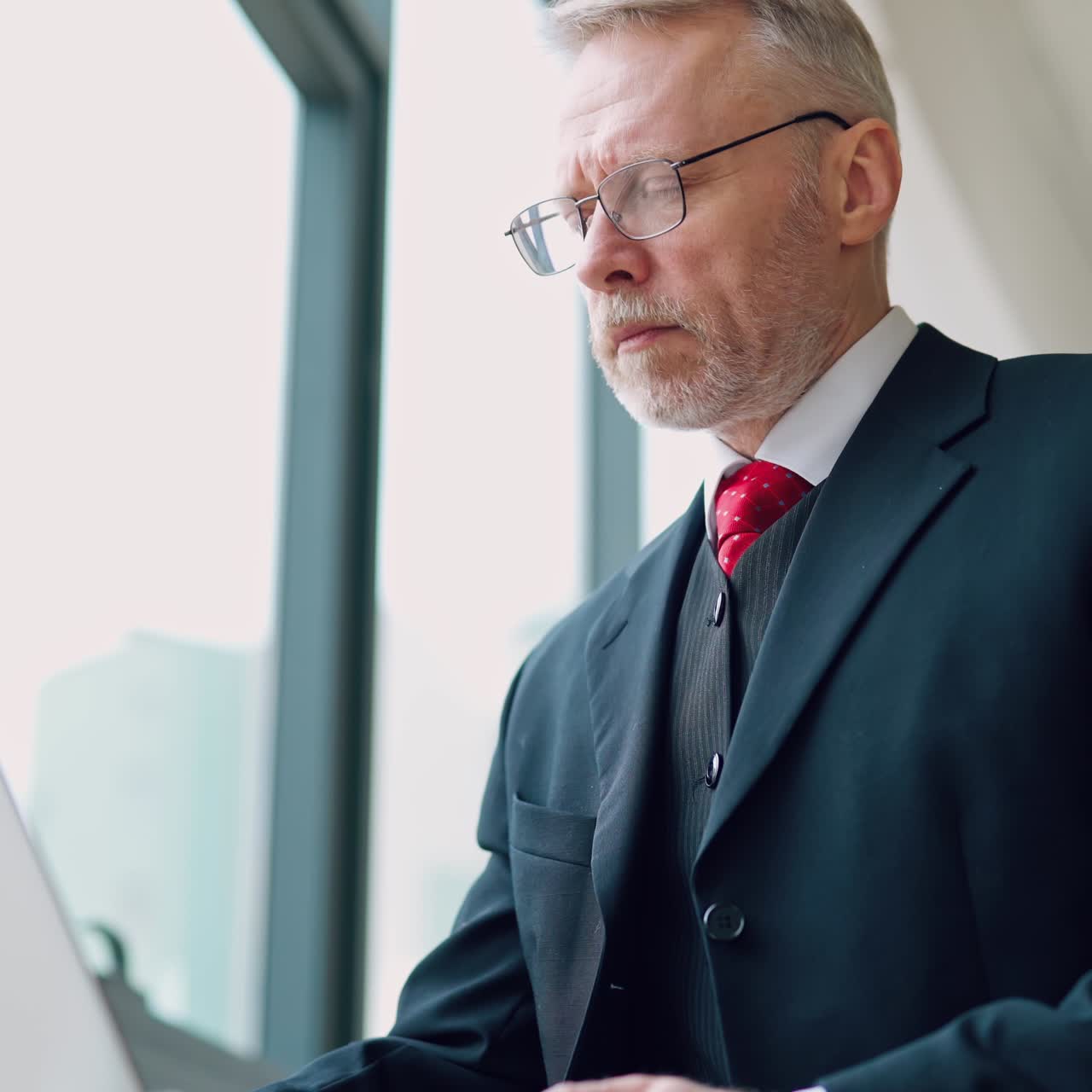 Senior serious businessman standing near window with panoramic city view. Laptop in hands, man thinking while checking e-mails standing near window. Video from the side.