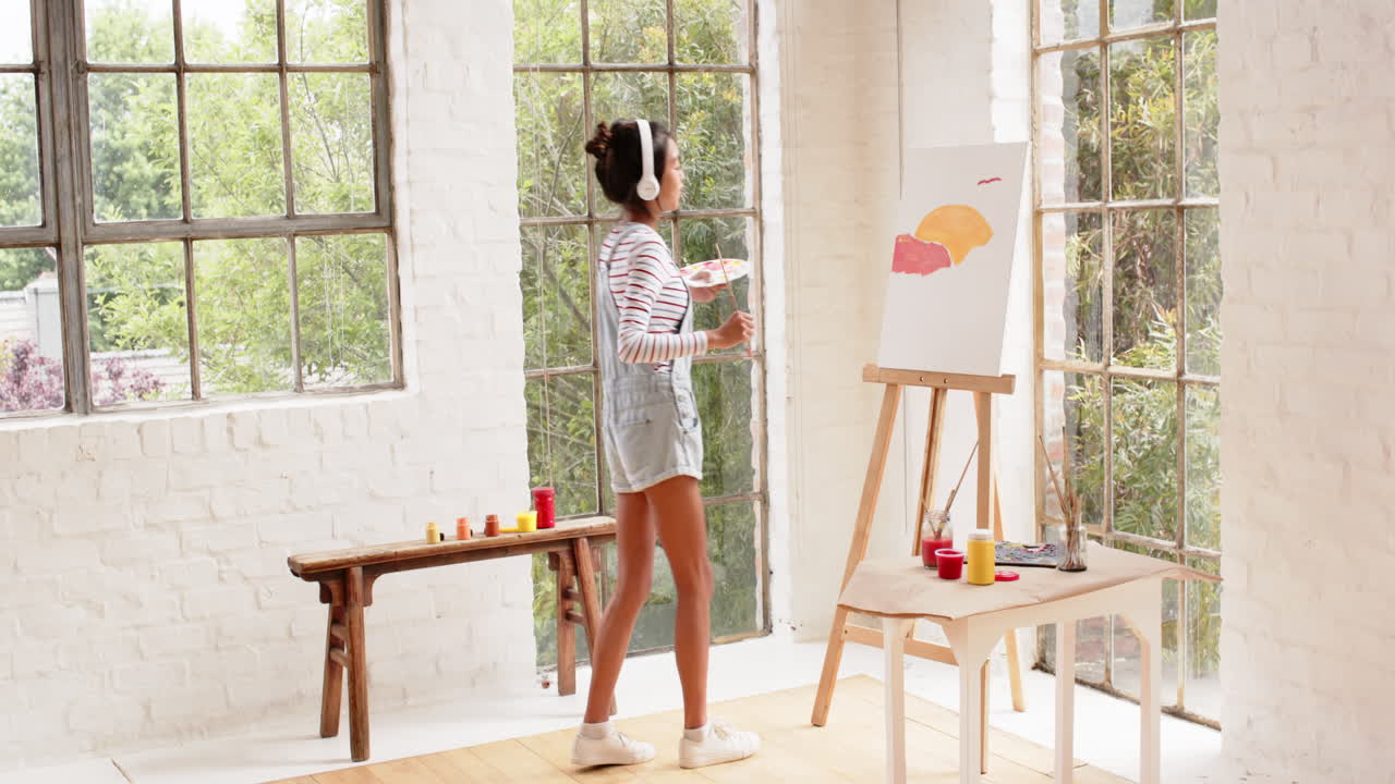 Asian young woman painting on canvas in bright studio, wearing headphones and overalls, at home