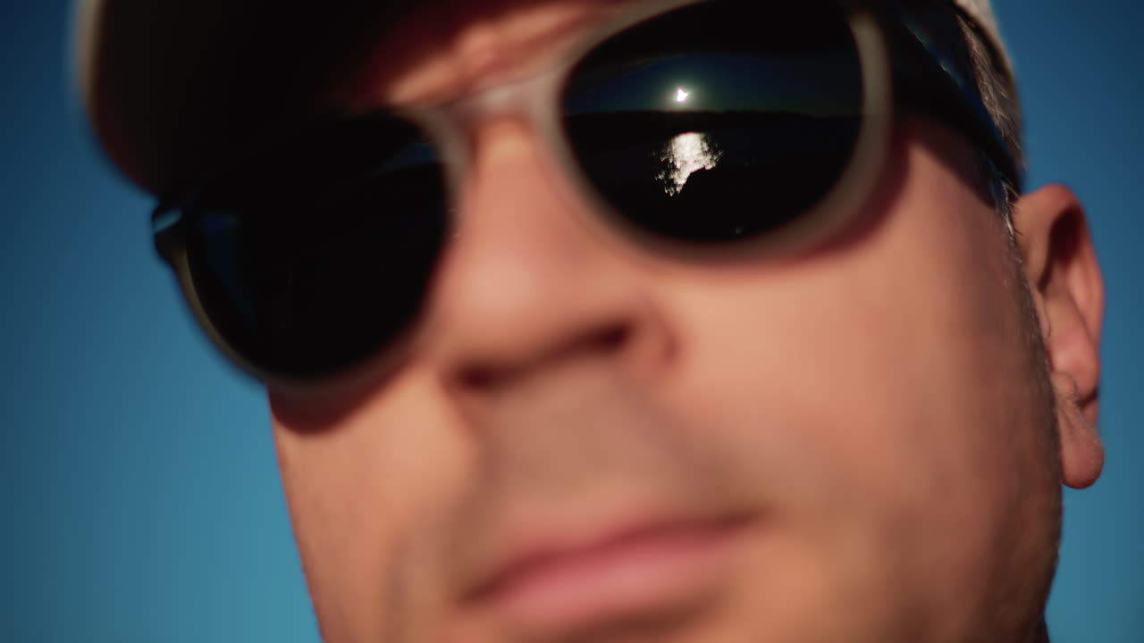 Extreme close up of a man wearing dark sunglasses and a cap, framed tightly against a clear blue sky in strong daylight