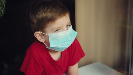 un niño con una máscara médica mira por la ventana 01