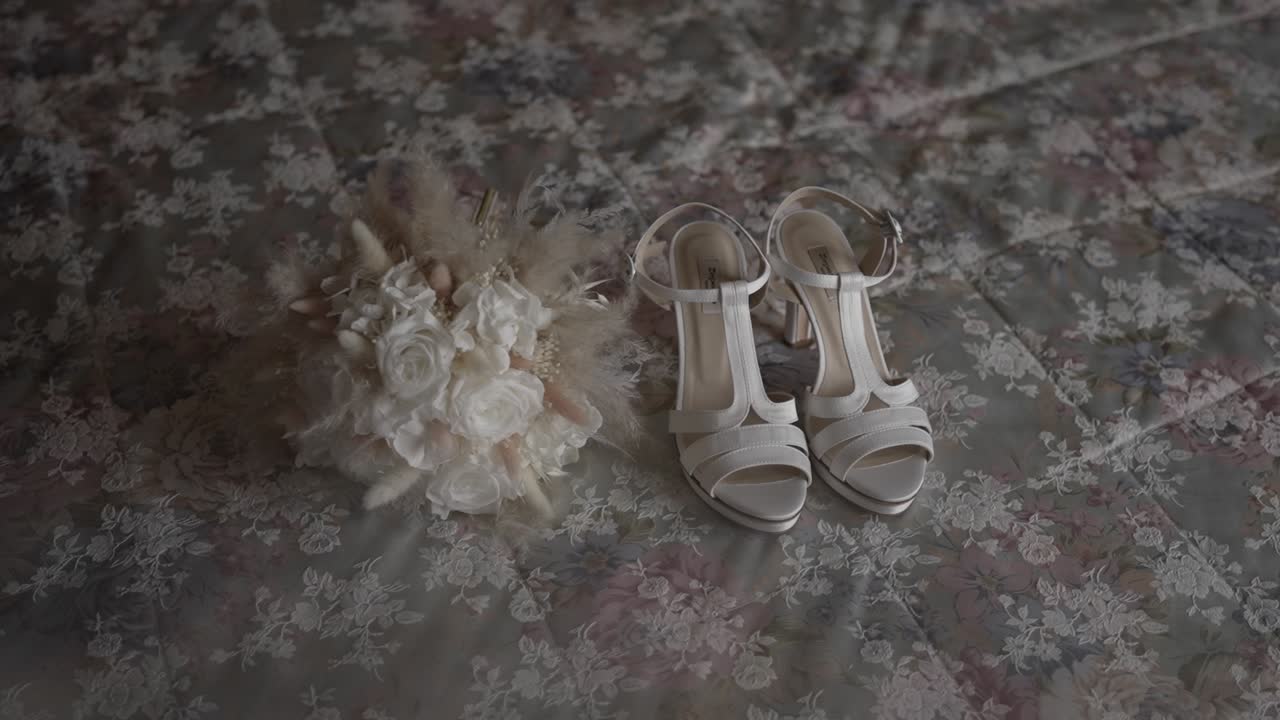 White bridal shoes paired with a delicate bouquet featuring white roses and soft accents, placed on a floral patterned fabric