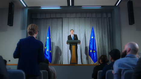 confident politician makes an announcement, answers media questions and gives interview. mature representative of the european union during performance at press conference. backdrop with eu flags.