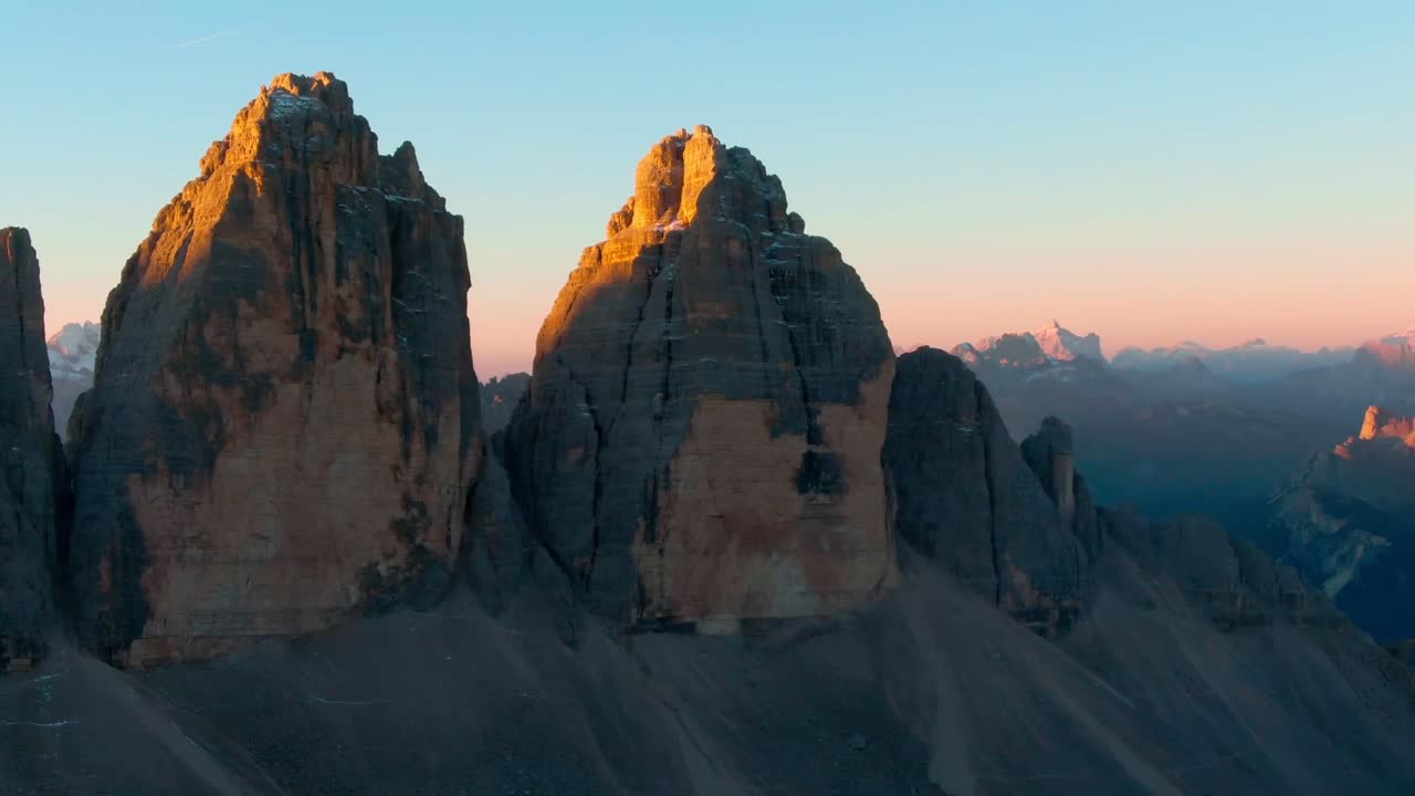 A drone panning shot of iconic Dolomite mountains being illuminated by first light from sunrise with distant jagged peaks in the distance lighting up under the golden glow.