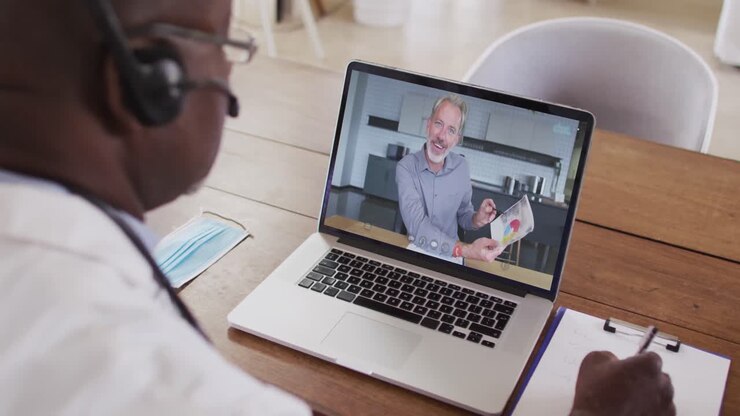 Afroamerikansk mandlig læge med headset på, der tager noter under et videoopkald på sin bærbare computer