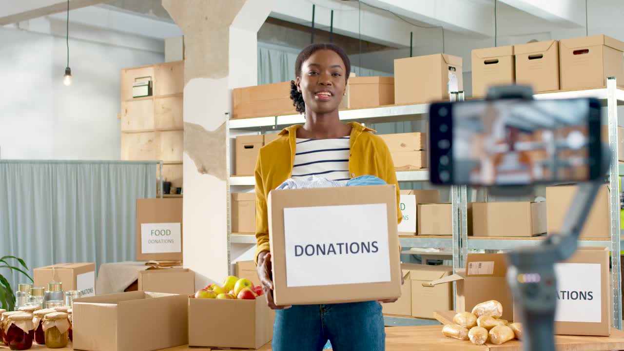 Young African American woman volunteer holding donation box and recording video blog on smartphone