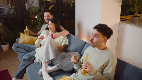 top view of a couple and a friend watching interesting movie on television sitting on couch, eating popcorn and chips and drinking soda