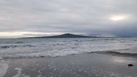 small waves rolling ashore with the magnificent rangitoto volcano in the background and dramatic clouds above