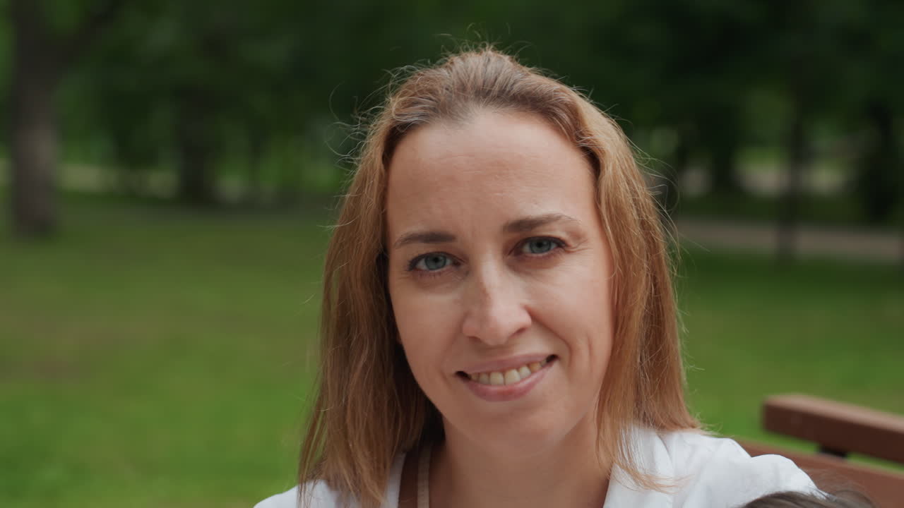 Madre e figlia sedute su una panchina, la madre sorride dolcemente alla telecamera, la testa della figlia parzialmente visibile, sfondo di parco illuminato dal sole con alberi verdi, camicetta bianca casual, espressione affettuosa, famiglia rilassata.