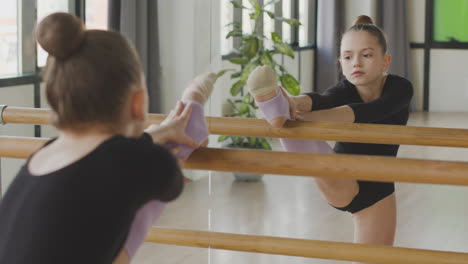 young ballerina practising leg positions at ballet bar with the help of her teacher