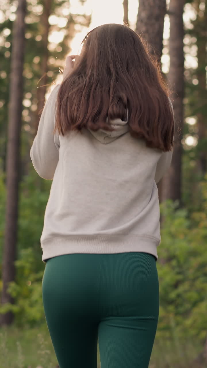 여성은 숲에서 달리는 헤드을 착용합니다. 일 후 조하는 젊은 여성은 일몰 때 자연에서 트랙을 듣습니다. 건강을 개선하기 위해 저녁 일상