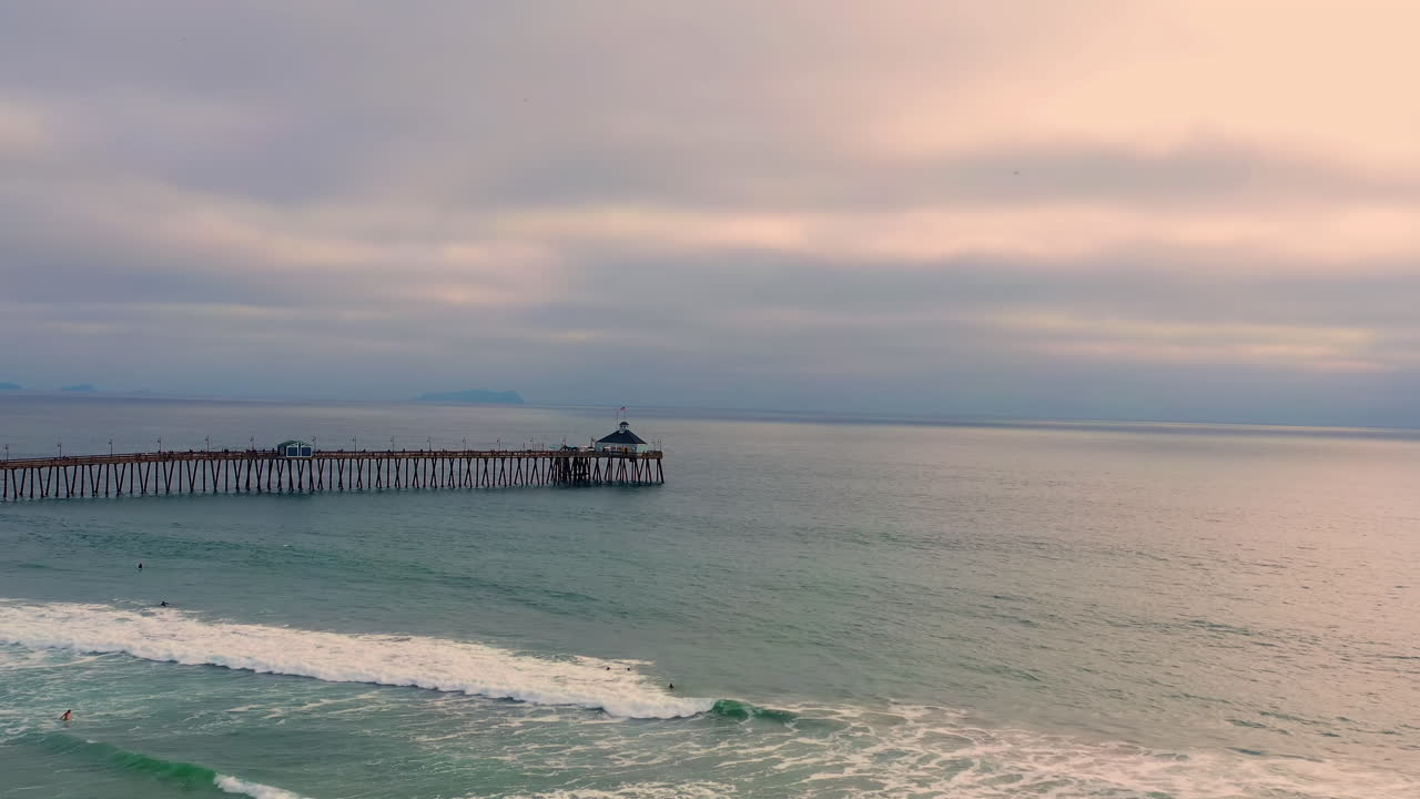 Drone Flies Toward Imperial Beach Pier Over Ocean Waves And Surfers In San Diego, California - aerial shot