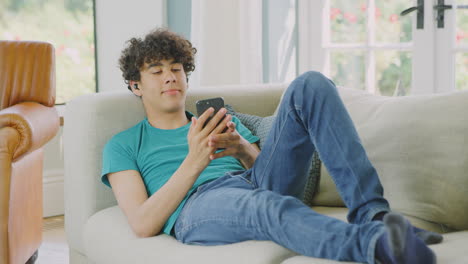 teenage boy wearing wireless earbuds posing for selfie on sofa at home using mobile phone