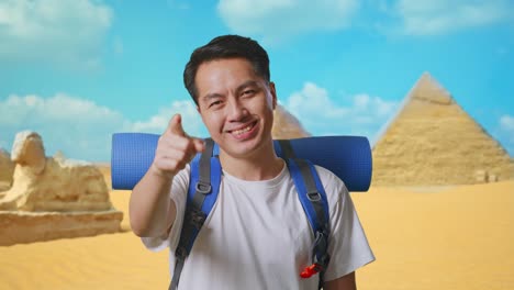 close up of asian male hiker with mountaineering backpack smiling, touching his chest, and pointing to camera while traveling in pyramid of giza