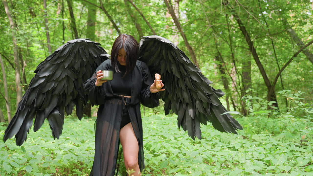 Mysterious black fairy creature with feathered wings walks through lush green forest holding snack and drink, gently drops metal cup on ground, blending elegance