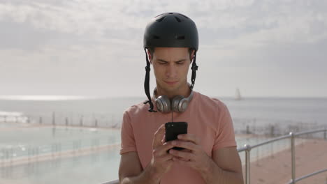 retrato de un joven guapo de pie junto a la playa enviando mensajes de texto usando un teléfono con casco