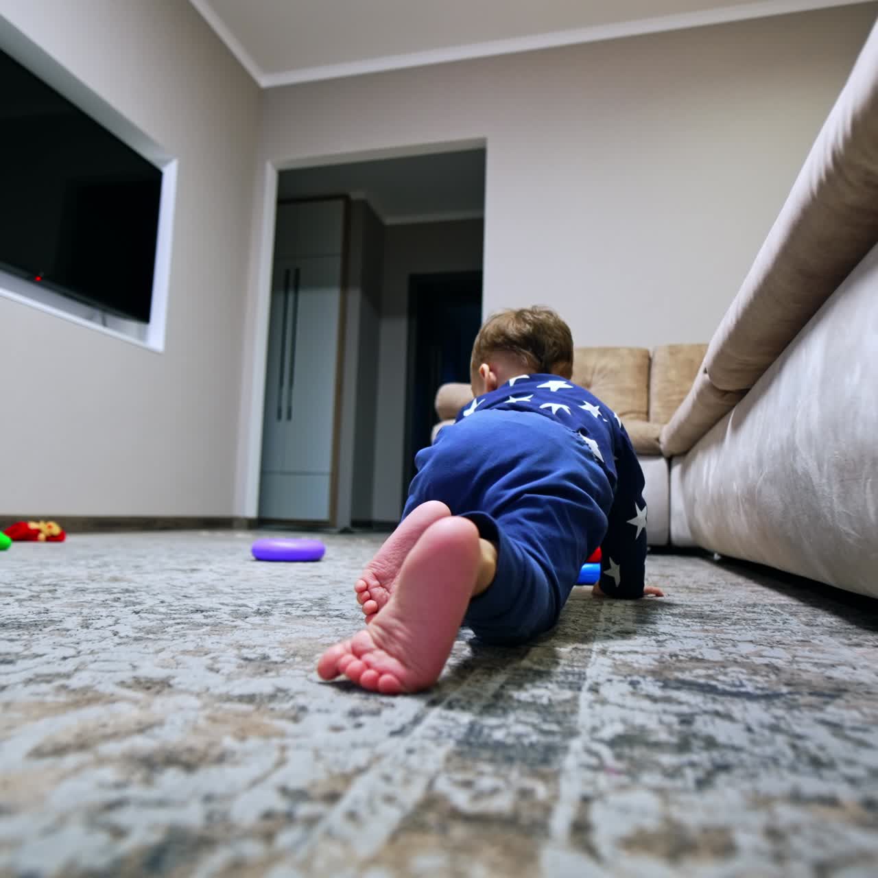 Dark-haired little boy in blue pajamas crawling by the carpet indoors. Toddler plays with a toy pyramid on the floor