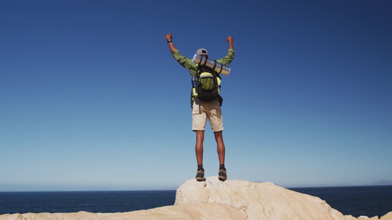 hombre afroamericano caminando en las montañas de pie en la roca levantando las manos por el mar
