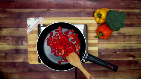 una vista superior de la adición de tomates en la sartén, colocados en una estufa, dos grandes cápsulas amarillas y rojas y un brócoli verde en la mesa