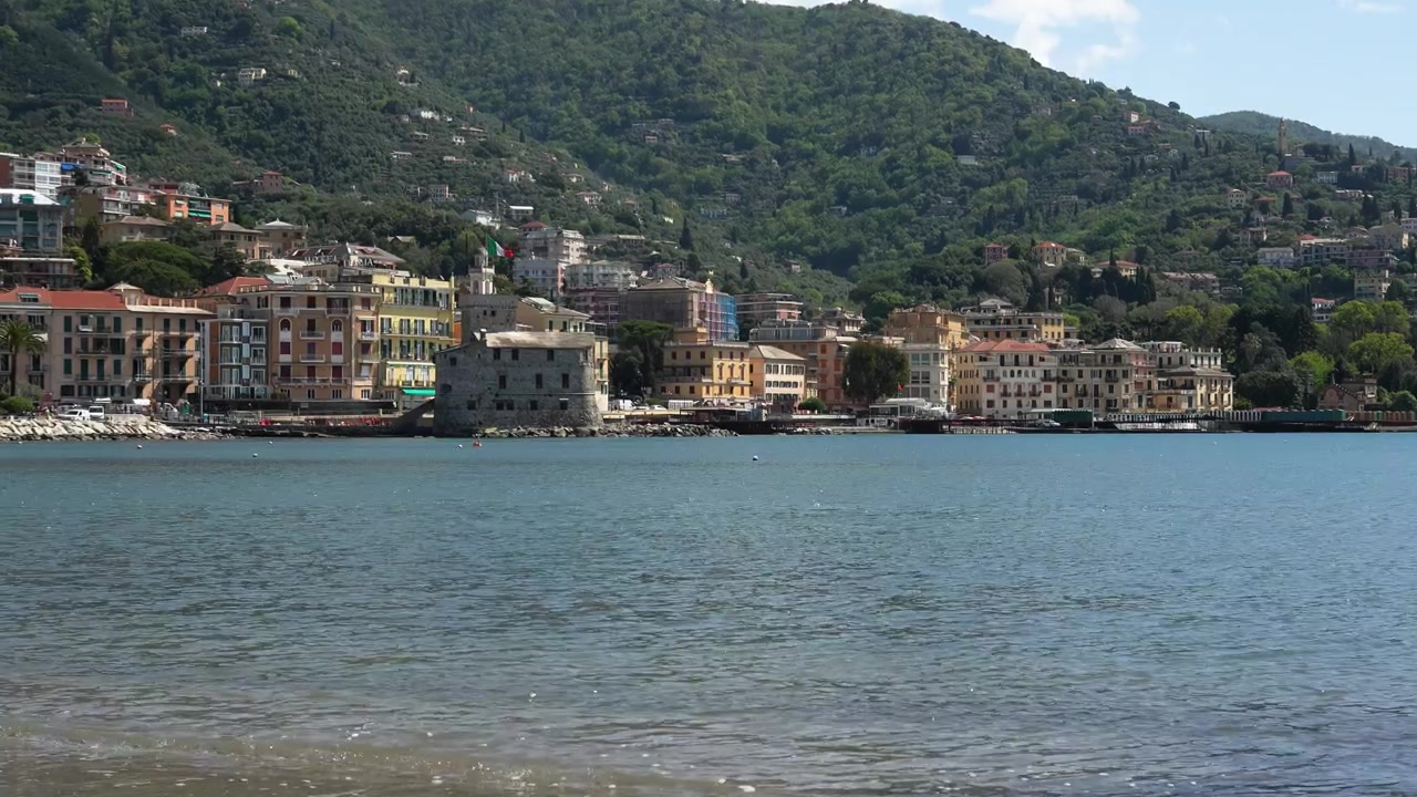 Inquadratura grandangolare di Rapallo, Italia, con gli edifici colorati sul lungomare e rigogliose colline verdi sullo sfondo, mare blu calmo in primo piano, piena luce diurna, atmosfera tranquilla, ripresa stabile, composizione vibrante.