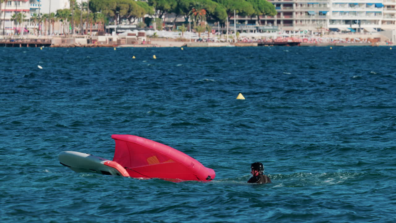 A man in a wetsuit falling down while wing foiling with a bright pink sail on the blue Mediterranean waters of Cannes, France