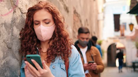 close-up view of redheaded woman wearing mask and using smartphone leaning on a wall in the street