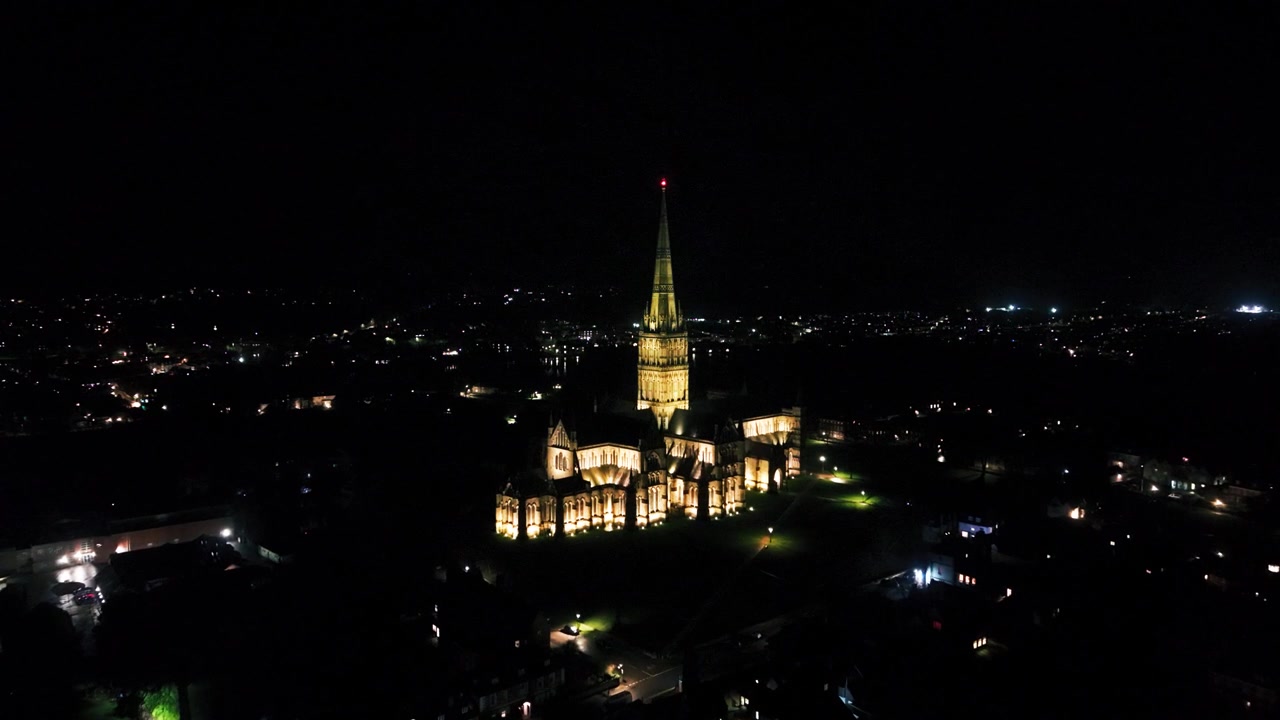 Wide aerial night view of Salisbury Cathedral illuminated against the dark cityscape, with its distinctive spire rising prominently above Salisbury in Wiltshire, England
