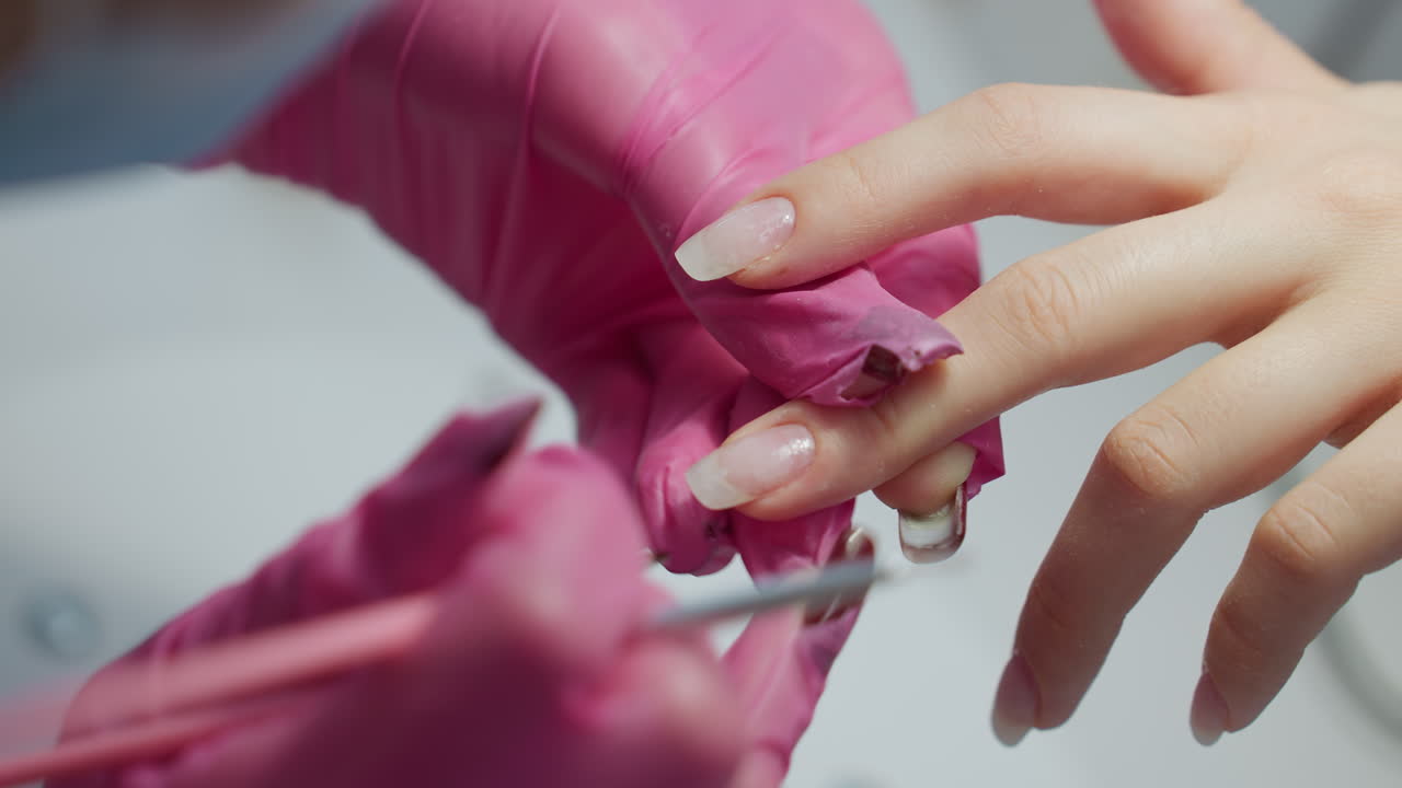 Close up view of nail technician in pink gloves switching between two brushes while working on client's fingernails, moving carefully to next finger during manicure session in clean beauty environment