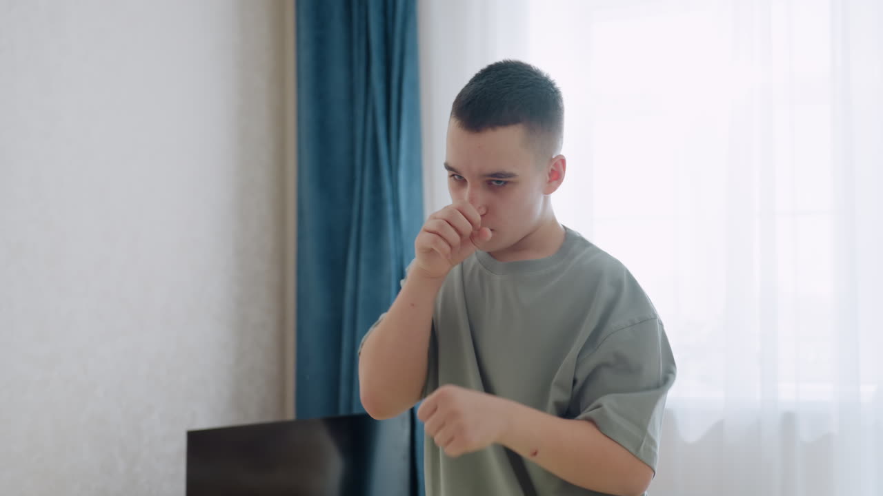 Teen boy with serious expression throws focused punch toward camera during indoor boxing practice session, wearing casual clothes near window with curtain
