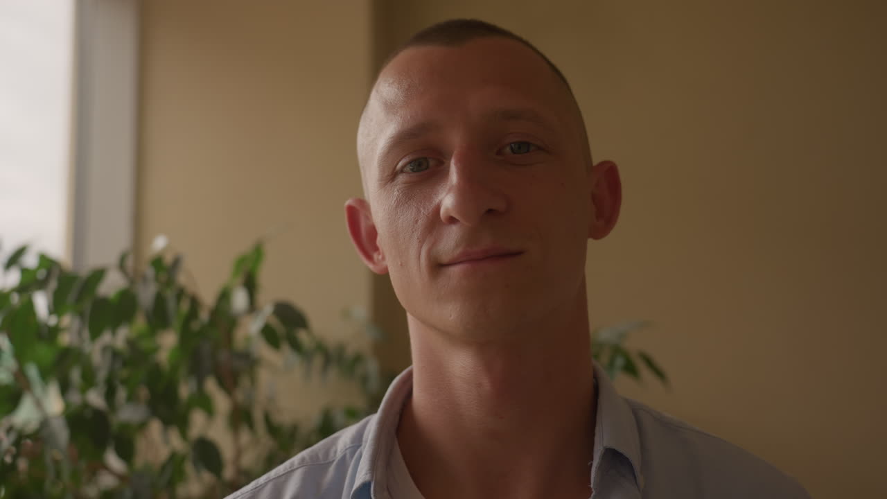 Uomo caucasico che guarda fuori dalla finestra, appartamento illuminato dal sole con pianta in vaso e vista sulla città, primi piani che rivelano un profilo introspettivo che si trasforma in un sorriso gentile, camicia casual e taglio buzz cut, caldo naturale