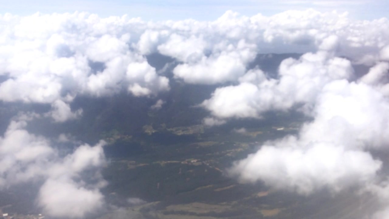 An arial view over puffy clouds with green land below.