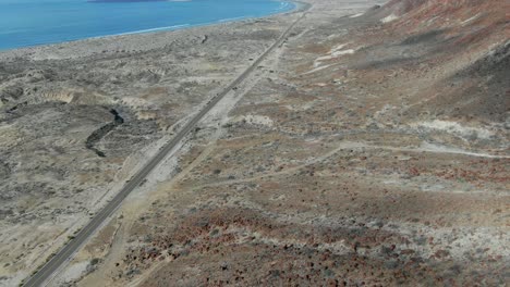 Aerial-view-tilting-up-shot,-Scenic-view-of-highway-besides-a-red-rocky-hill-in-La-Purisima-Baja-California-sur,-Mexico,-blue-sea-in-the-background