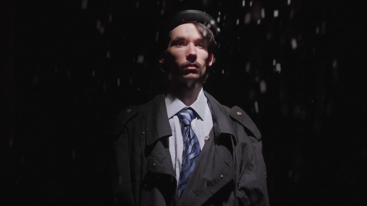 A lone young man in a dark coat and hat stands centered against a black background, illuminated by a narrow overhead spotlight with rain-like streaks falling