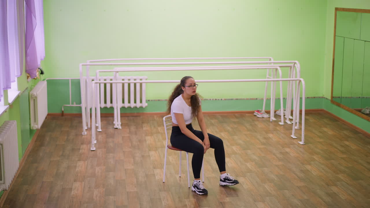 Lady in white top and black leggings sinks onto chair looking stressed after workout in bright studio, wooden floor and mirror bars behind, capturing fatigue, pause, breath