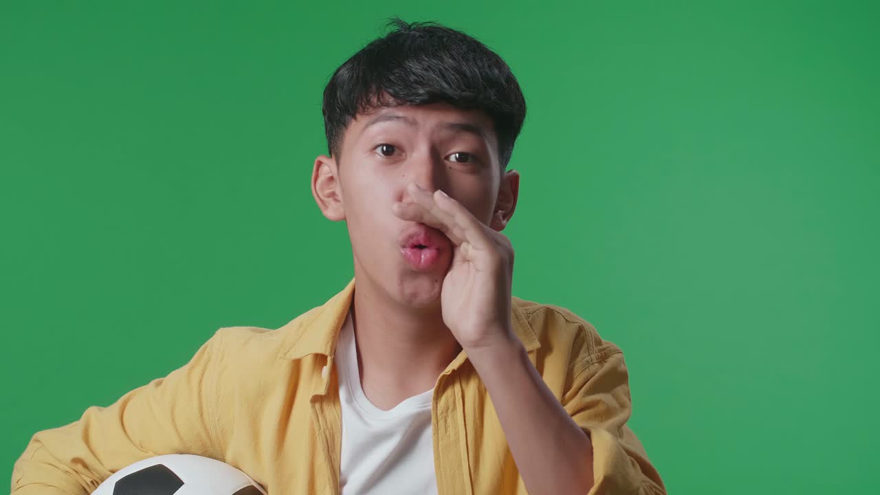 초록색 스크린 배경에서 축구 경기를 응원하는 동안 패자를 조롱하는 공을 가진 아시아 소년의 클로즈업