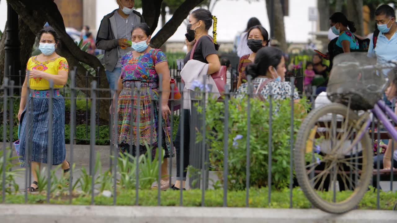 Antigua Guatemala three native indigenous women with traditional dresses walk along a busy town square wearing face masks due to pandemic along with various pedestrians - Handheld slow motion