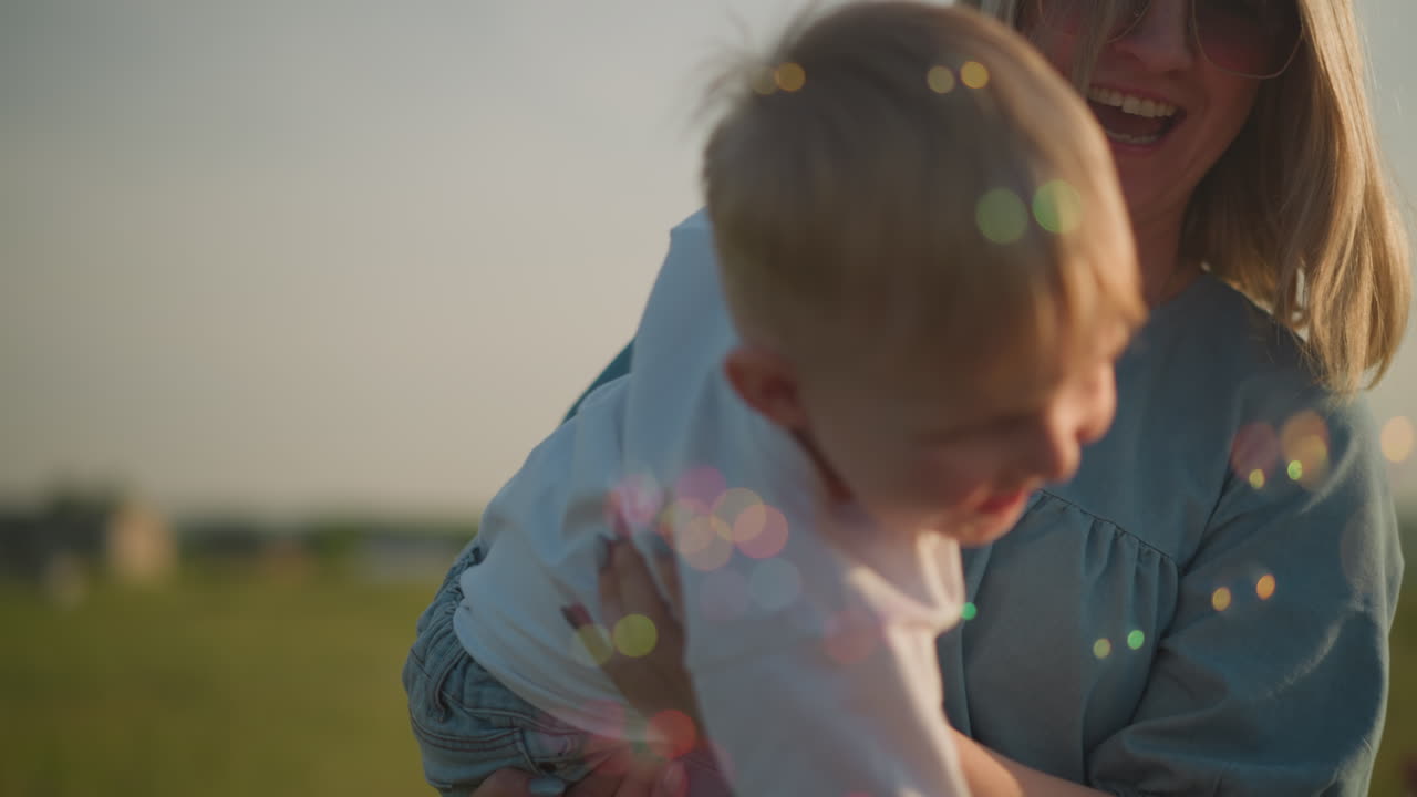A happy woman in a blue dress holds a little boy wearing a white shirt, both laughing as they enjoy colorful bubbles floating around them in a grassy field, creating a heartwarming and playful moment
