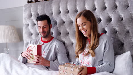 Mujer-Abriendo-Un-Regalo-De-Collar-En-La-Cama-En-Casa-Mientras-Una-Pareja-Intercambia-Regalos-El-Día-De-Navidad