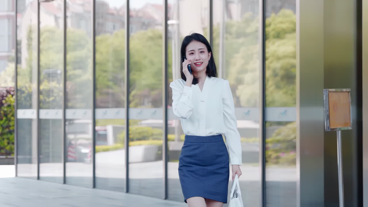 开朗的年轻职业女商人自信地接听电话，走在明亮的玻璃办公室走廊空间里，这里拥有全景城市景观和现代室内设计。