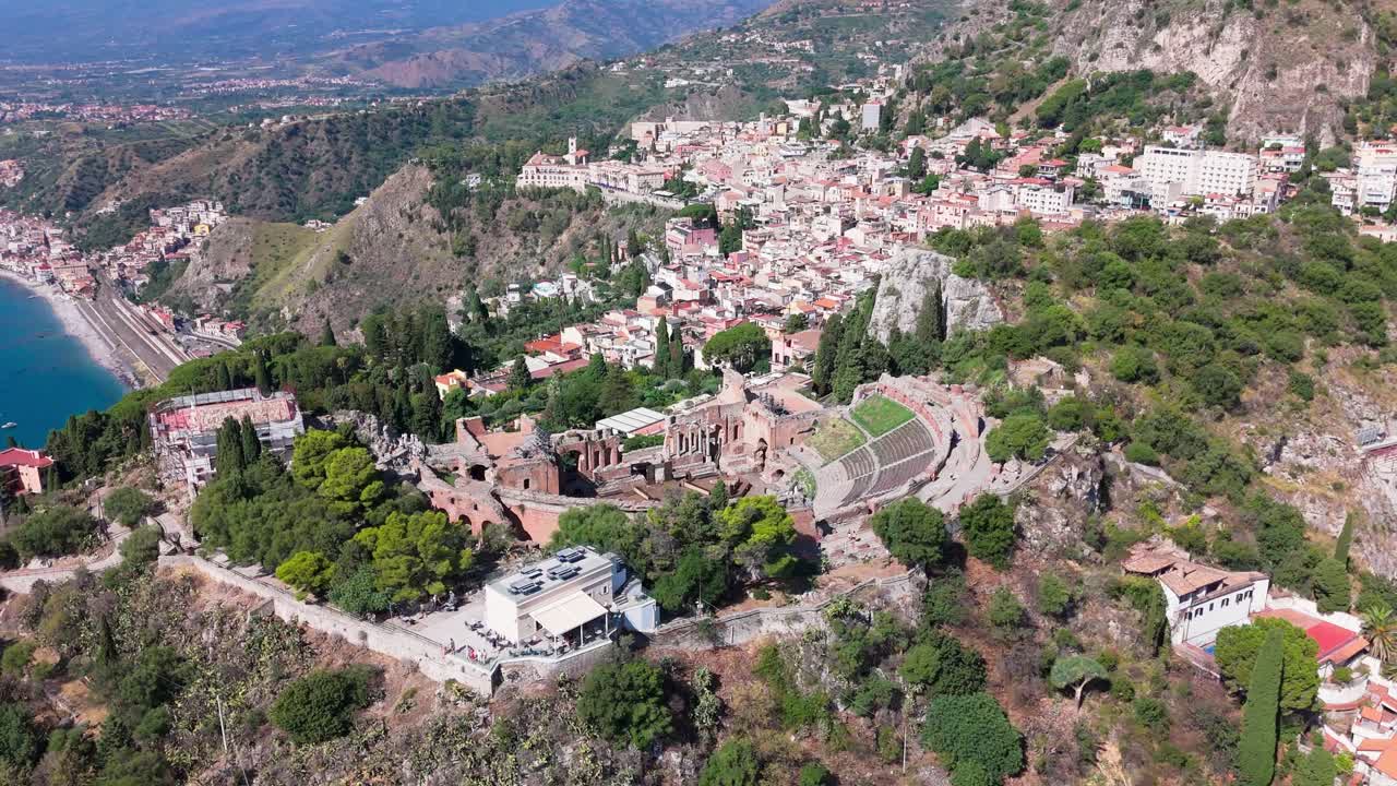 Drone slowly orbits the amphitheatre from left to right, revealing the historic site with the town and coastline in the background