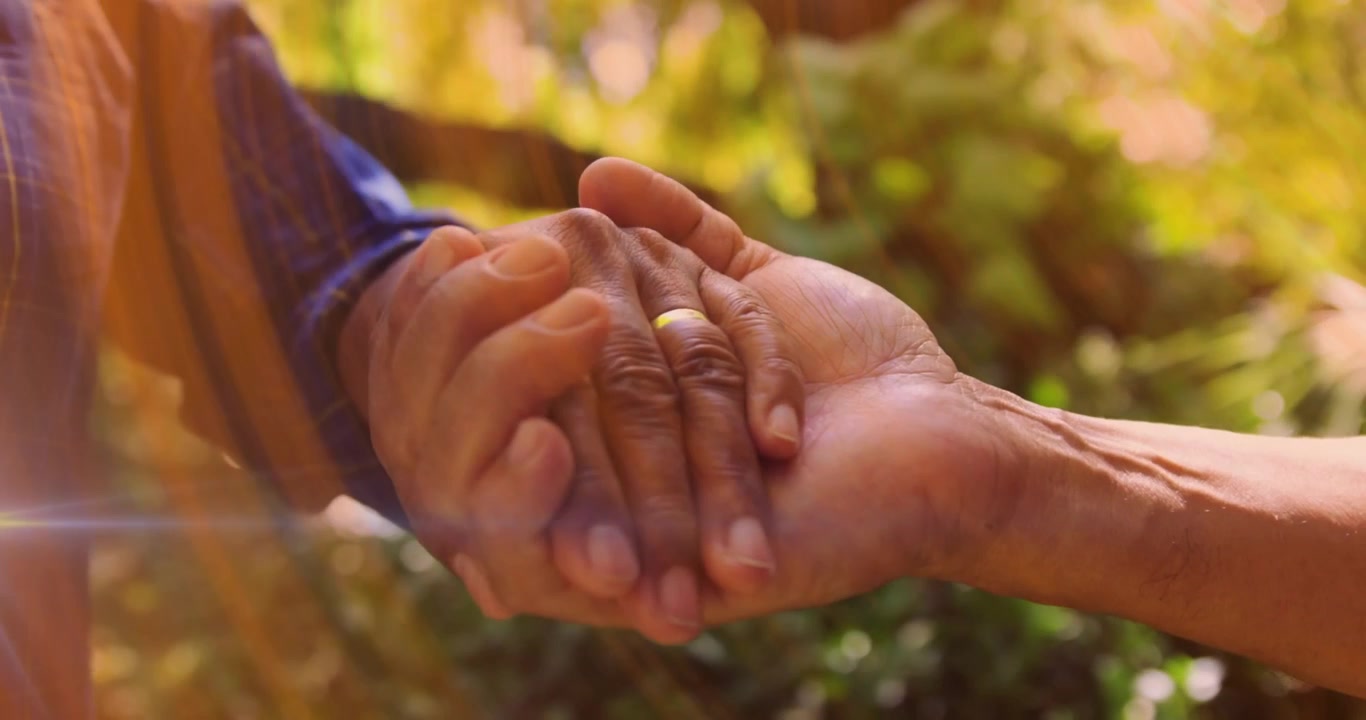 Pareja mayor tomándose de las manos en un jardín soleado, entrelazando los dedos sobre la alianza de oro para la salud.