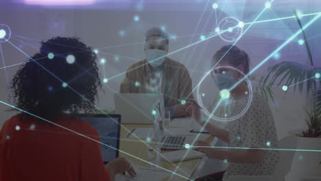 animation of glowing network over diverse colleagues in face masks using computers in office