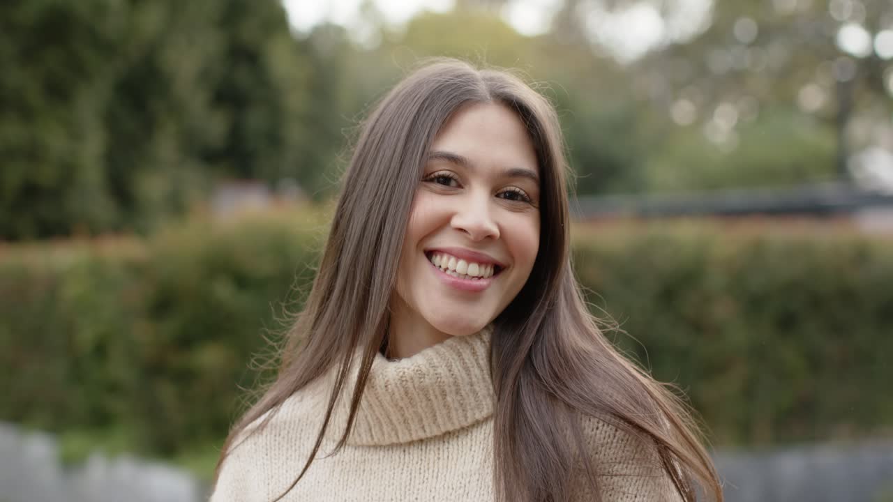 Mujer con suéter de punto protegiéndose los ojos, cepillándose el cabello y sonriendo en el jardín con un reloj inteligente.