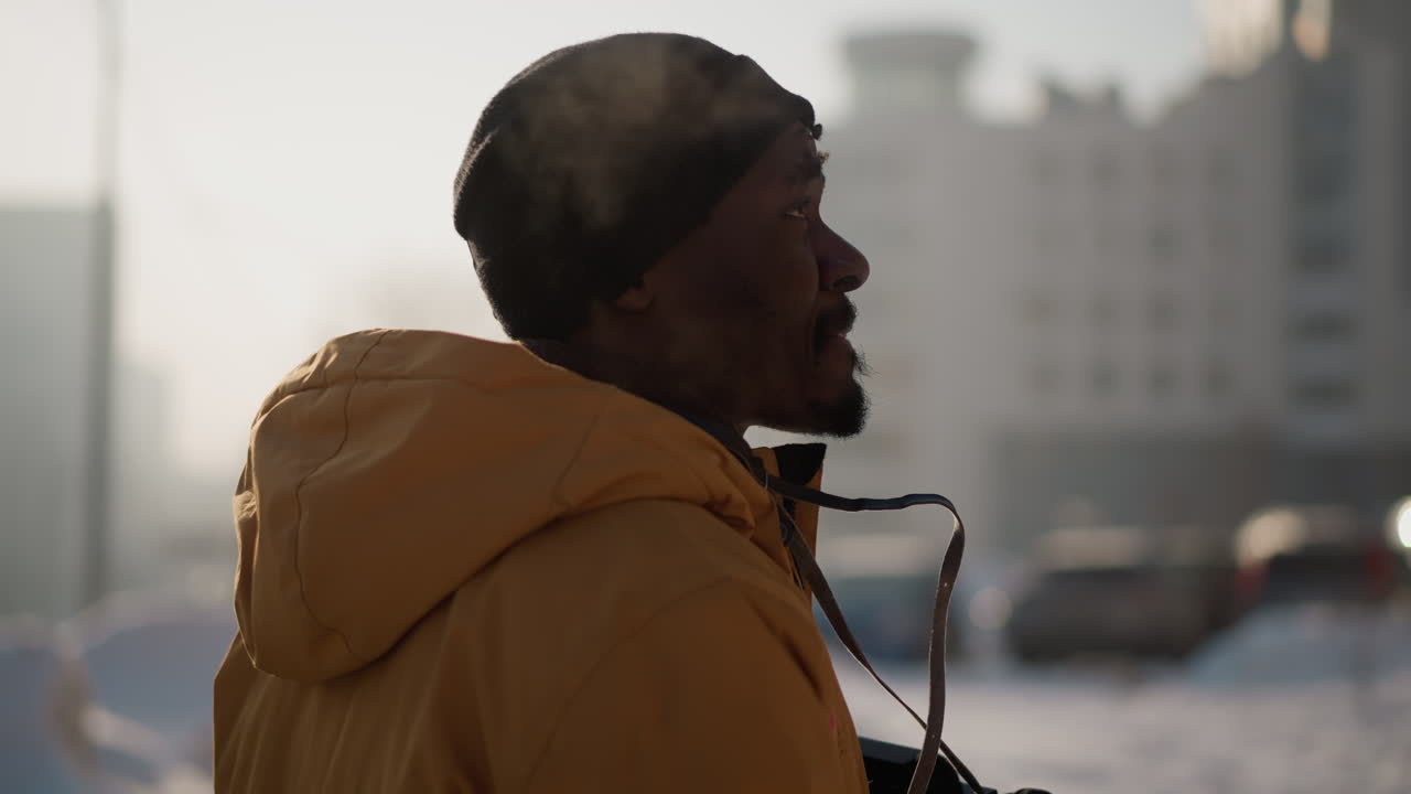 back view cinematographer wearing insulated winter coat walking along snow covered urban sidewalk flanked by parked cars tall buildings while exhaling visible breath in crisp frosty morning air