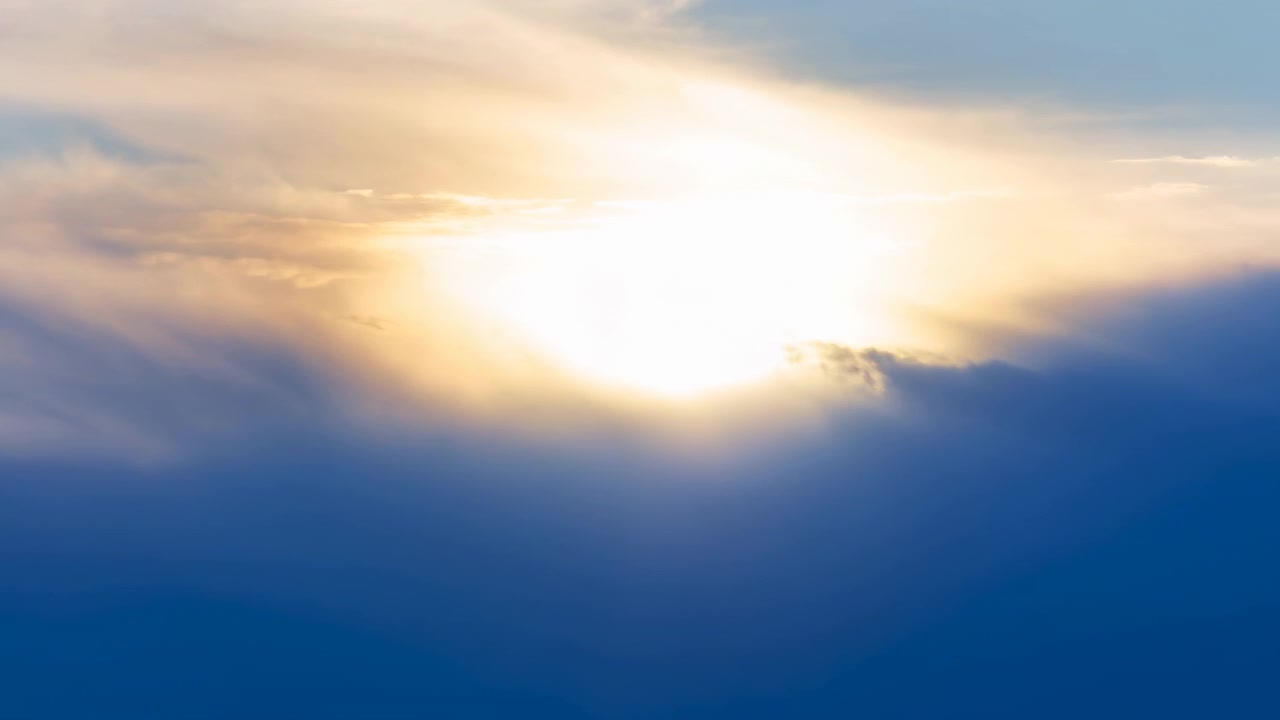 Low sun remains partly veiled by drifting cloud layers while broad beams of warm light fan outward, illuminating pale upper clouds above a deep blue lower cloud bank.