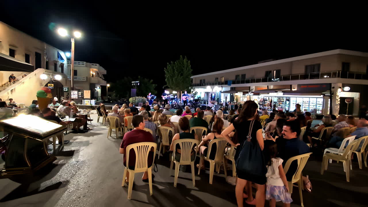 Profile view of Greek locals and tourists enjoying concert going on at Agios Nikolaos during nighttime.