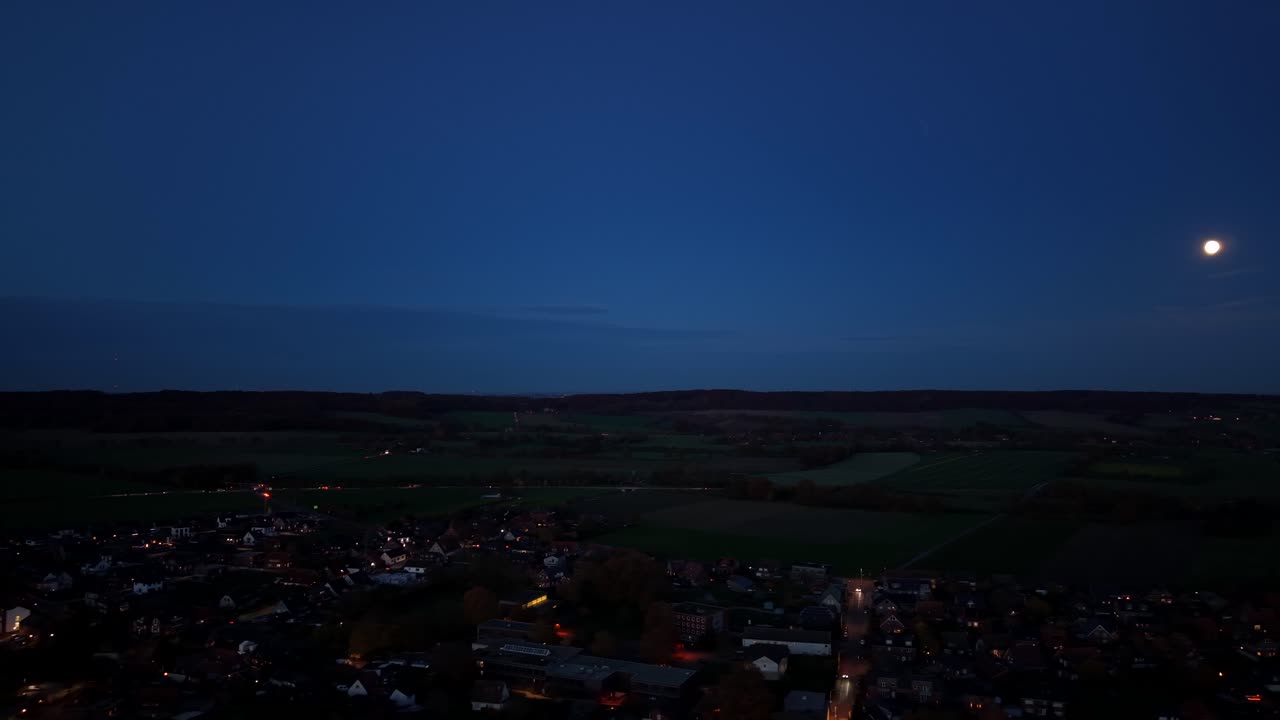 Full moon at blue dusk sky in american town. Lighting houses and streets in USA. Aerial lateral wide shot. Rural landscape with hill farm fields at night. Panorama view