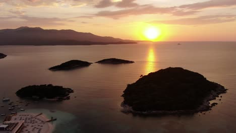 drone view in albania flying over blue crystal clear water on sunset, with the sun setting in the horizon with small islands in ksamil