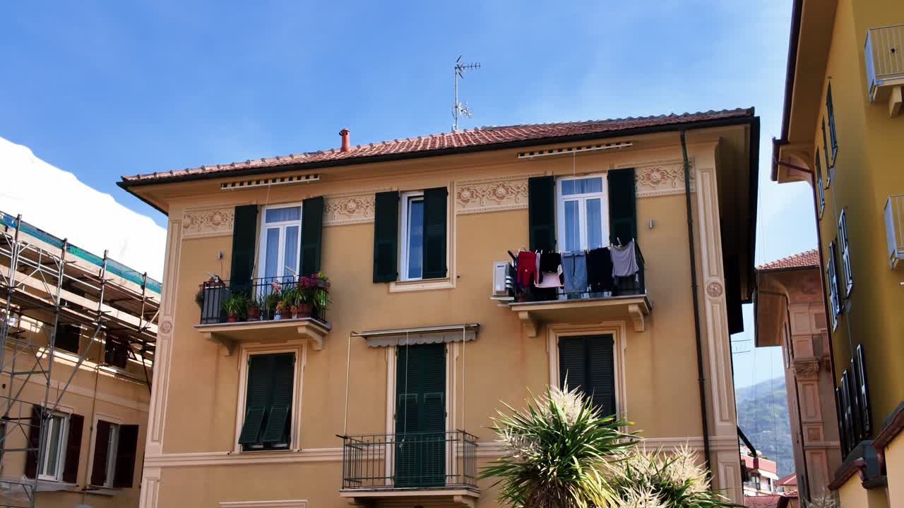Luz solar quente ilumina charmosas fachadas de apartamentos mediterrâneos amarelos com persianas verdes, varandas exibindo plantas e roupa estendida, e palmeiras. Sem pessoas, plano estático de grande angular.