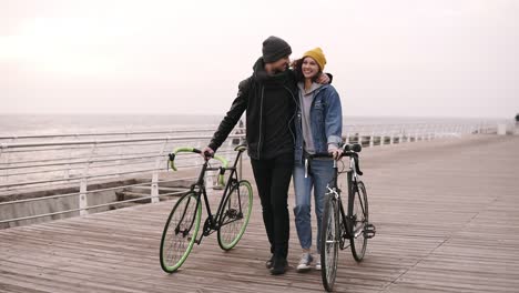 Full-Length-Of-Couple-Of-Young-Hipsters-Walking-Together-With-Their-Bikes-Near-The-Sea-At-Autumn-Day