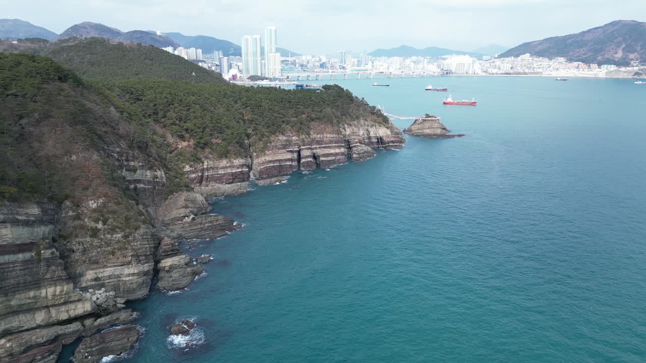 Drone aerial view in South Korea countryside flying over the clear blue sea of Busan next to a green mountain, ships and city in the background sunny day